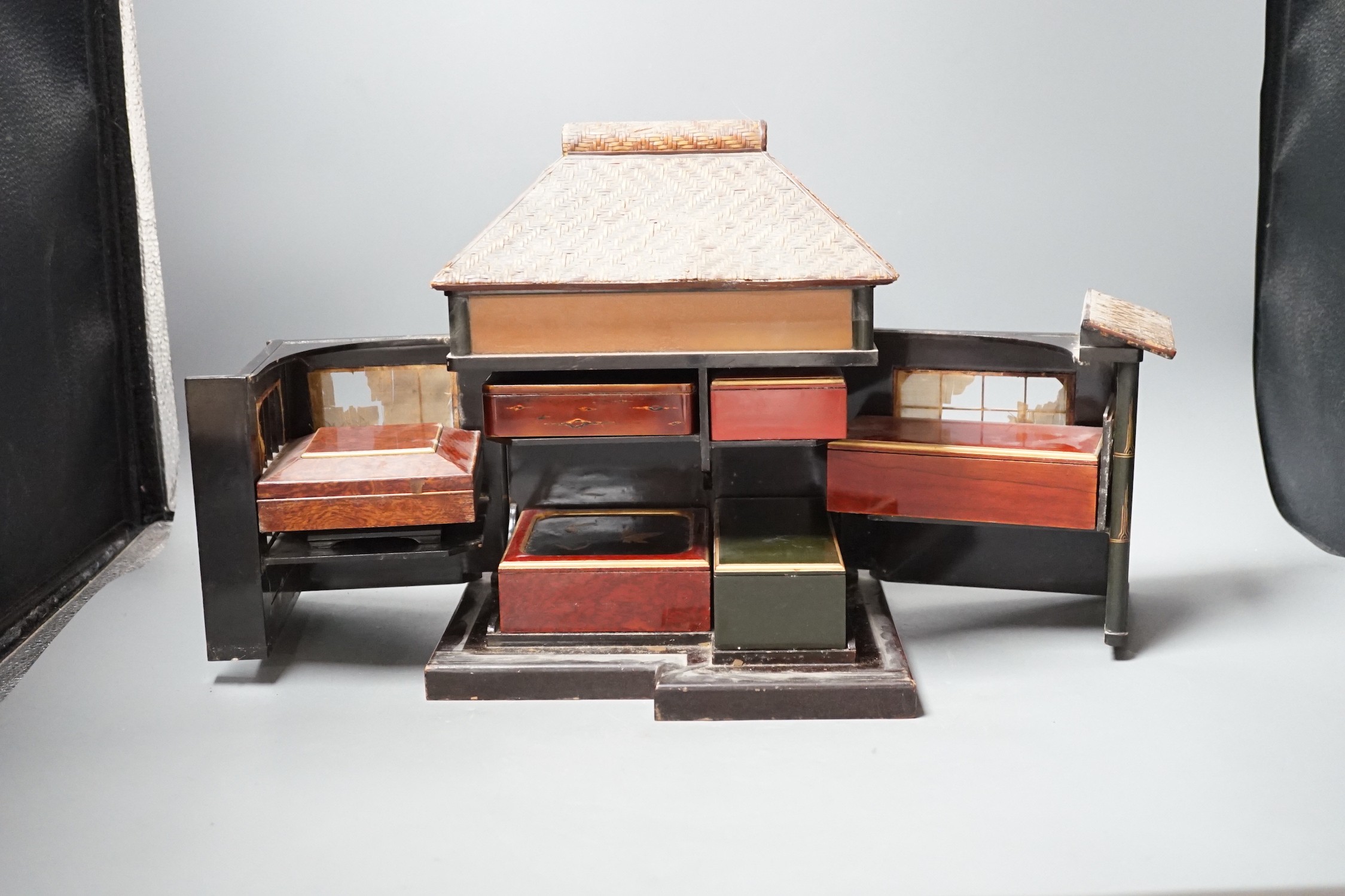 A Japanese lacquer 'house' with pitched wicker roof and opening side and front sections revealing internal shelves and six lacquer boxes and a small glass painted panel, ‘house’ height 28.5 cms high